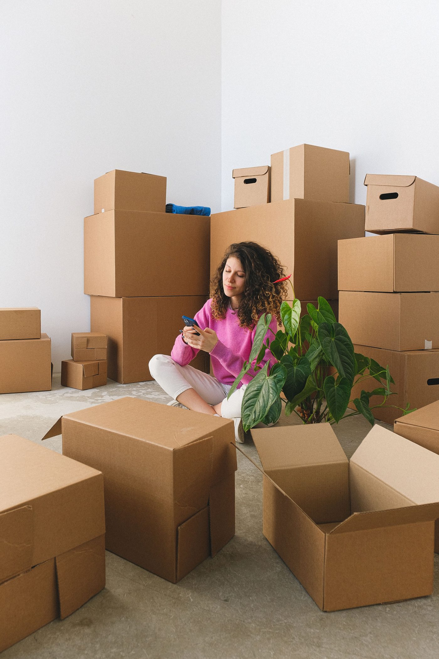 A person sitting on the floor surrounded by stacked cardboard boxes, checking their phone after a move.